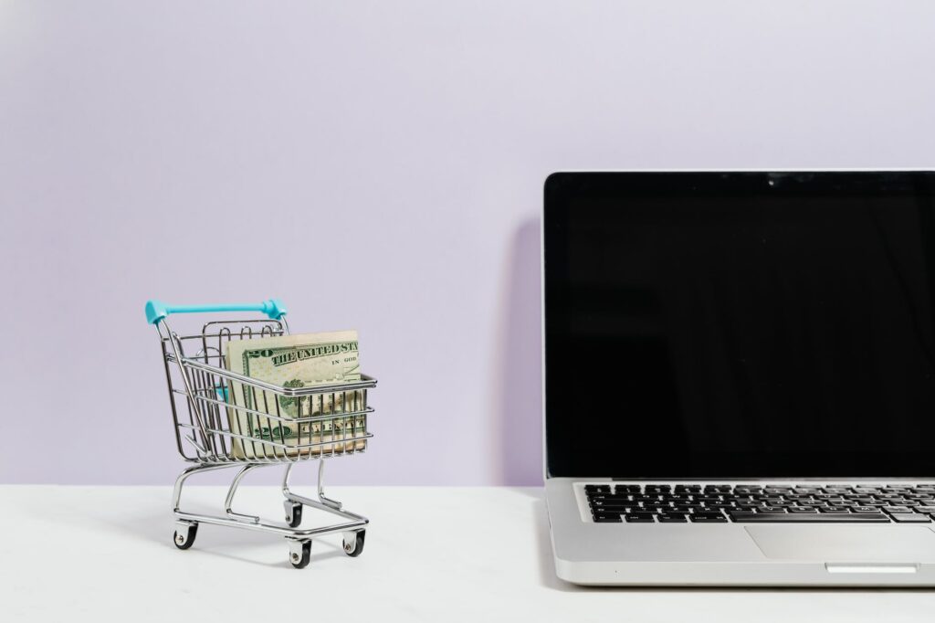 macbook pro on white table beside a miniature shopping cart with money